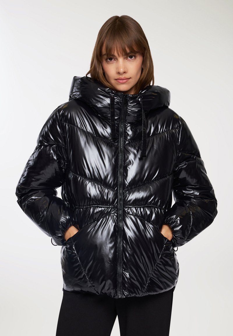 Giacca a vento nera con superficie lucida e trapuntata, cappuccio oversize, zip frontale e due tasche laterali; presenta un design con vita arricciata.