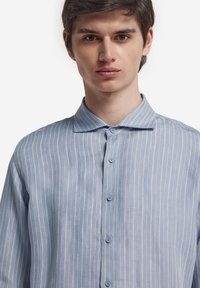 Ljusblå skjorta med knappar och tunna vita ränder, med standardkrage och blå knappar. Tyget ser jämnt och lätt ut.