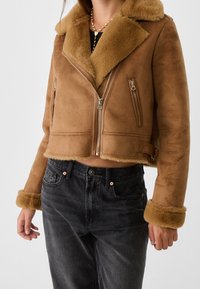 Giacca cropped in shearling marrone con colletto e polsini in pelliccia, tasche con zip e una texture esterna liscia. Abbinata a jeans scuri.