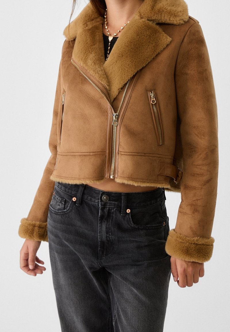 Giacca cropped in shearling marrone con colletto e polsini in pelliccia, tasche con zip e una texture esterna liscia. Abbinata a jeans scuri.