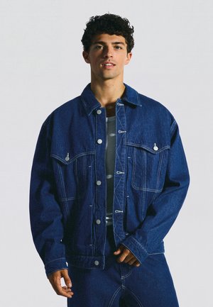 Ung mand iført en blå denimjakke og matchende jeans med et neutralt ansigtsudtryk.