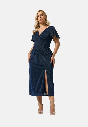 Femme portant une robe midi bleu foncé pailletée avec des manches volantées et une fente à l'avant, associée à des talons argentés à bride cheville, debout devant un fond uni.