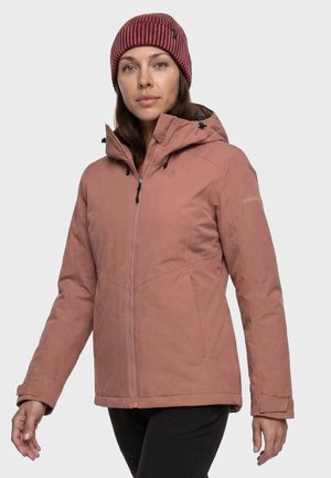 Junge Frau trägt eine staubrosa Kapuzenjacke und eine burgunderrote gestrickte Mütze, steht vor einem schlichten hellen Hintergrund.