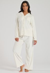 Witte pyjama-set van zachte stof, met een knoopsluiting bovenkant met een zak en lange mouwen, gecombineerd met wijde broekspijpen.