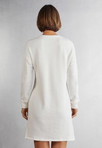 Vestido de suéter blanco con canalé y mangas largas, presenta un corte relajado y sutiles cortes en los lados del dobladillo para facilitar el movimiento.