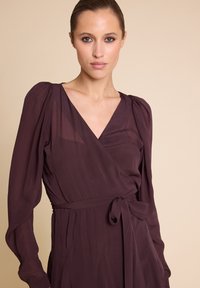 Mørk lilla omslagskjole med lange pufferm, v-hals og en knyttet midje detalj. Myk, flytende stoff med en glatt tekstur.