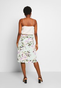 Jupe midi florale en tissu vert menthe avec un motif de fleurs violettes et blanches, associée à un débardeur de couleur claire et des sandales à talons carrés.