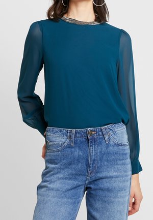 Woman wearing a teal long-sleeve blouse with sheer sleeves and metallic neckline paired with high-waisted light blue jeans.