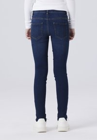 Tumši zili skinny džinsi ar gludu tekstūru, piecu kabatu dizainu un baltām kedām, skatīts no aizmugures.