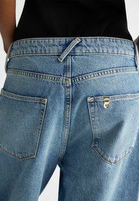 Takakuva henkilöstä, joka päällä vaaleansiniset farkut, joissa on suuret takataskut, ja musta paita on sisään työnnetty.