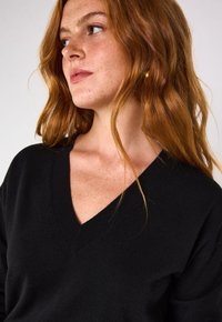 Maglione nero a scollo a V con una consistenza morbida e collo a costine, indossato da una persona con capelli rossi mossi, che mette in evidenza una vestibilità rilassata.