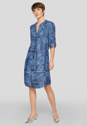 Frau mit kurzen Haaren, die ein blaues knielanges Kleid mit Blumen- und Schriftmustern trägt, steht mit einer Hand an der Seite und silbernen Absätzen.
