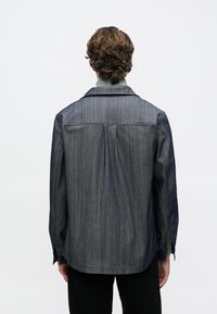 Szemüveges, göndör barna hajú személy sötétkék farmerdzsekiben és világosszürke garbóban, háttal áll egyszerű háttér előtt.