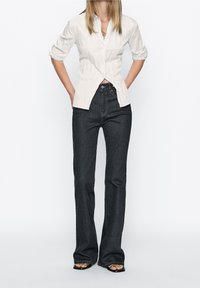 White button-up shirt with three-quarter sleeves, paired with dark blue flared jeans, showcased with black open-toe sandals.