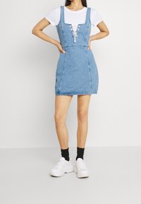 Denimimekko, jossa on tiukka muotoilu, olkaimet ja neliökaula-aukko, nauhat edessä ja mini-pituus. Päällä valkoinen t-paita.