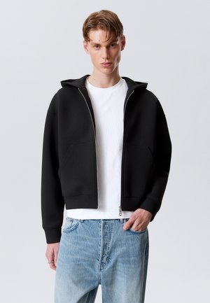 Black zip-up hoodie made of smooth fabric, featuring a hood, two front pockets, and ribbed cuffs, paired with light blue jeans.