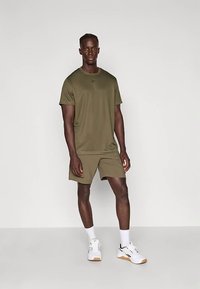 Olive krāsas sporta apģērbs, kas sastāv no īsām piedurknēm t-kreklu un šortiem, izgatavots no viegla auduma, apvienots ar baltām sporta kurpēm.