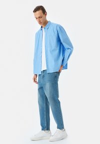 Camisa de rayas azul claro con mangas largas, cierre frontal de botones y un bolsillo, combinada con jeans azul claro y zapatillas blancas.