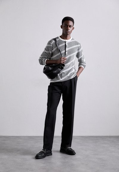 Μαύρος και άσπρος ριγωτός σκούφος, μαύρο παντελόνι και μαύρα παπούτσια. Κρατάει μια μαύρη τσάντα; στέκεται μπροστά σε μια ουδέτερη φόντο.