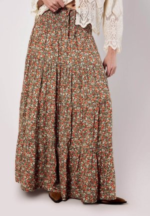 Person iført et langt, flerlaget blomsterprintede nederdel i orange, hvid og grøn, kombineret med en cremefarvet, cropped blonder top og brune spidse sko.