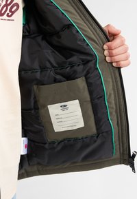 Interiorul jachetei cu umplutură este de culoare neagră, cu detalii verzi și o pungă din material canvas. Punga afișează eticheta de îngrijire și detalii de înregistrare. Materialul este nylon.