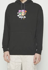 Personne portant un sweat à capuche noir avec un personnage de dessin animé tenant un ballon de basket et une balle de tennis sur la poitrine, assorti à un pantalon cargo beige.