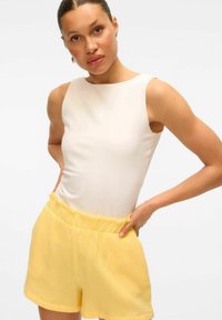 Biała bluzka bez rękawów w połączeniu z teksturowanymi żółtymi szortami z falbanką w pasie. Materiał jest miękki i ma lekką, casualową estetykę.