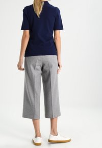 Persona con codino biondo che indossa una maglietta blu navy a maniche corte, pantaloni cropped grigi a gamba larga e sneakers bianche con suola in gomma, di spalle.