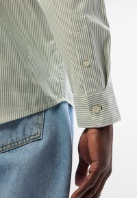 Πράσινο και λευκό ριγέ πουκάμισο με κουμπιά στους καρπούς και τσέπη. Φορεμένο με ανοιχτό μπλε τζιν που έχει αντιθετική κίτρινη ραφή.