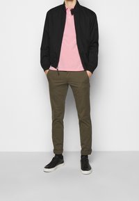 Zwarte bomberjack, roze polo, olijfgroene chino's en zwarte sneakers. De outfit heeft een gladde stofafwerking en een relaxte pasvorm.