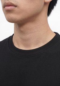 Nahaufnahme des unteren Gesichts und Halses einer Person, die ein einfaches schwarzes Rundhals-Shirt vor weißem Hintergrund trägt.