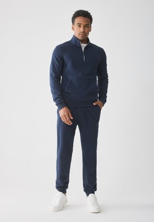 Man staat in een marineblauwe ritstrui en bijpassende joggers met witte sneakers tegen een effen witte achtergrond.