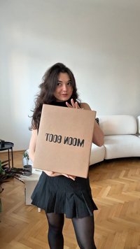 Boîte en carton brun avec texte noir "WOOM BOOT" tenue à la verticale. La boîte présente des surfaces lisses et sans ornements. En arrière-plan, un canapé clair et des plantes.