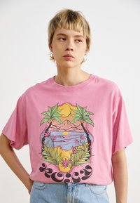 Pink cotton T-shirt featuring a tropical graphic design with palm trees, mountains, and a sunset, accented with bold black lettering.