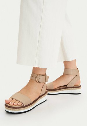 Pies con sandalias de plataforma beige con pulsera en el tobillo y suelas blancas y marrones, combinadas con pantalones de pierna ancha y cortos de color blanco roto sobre un fondo blanco.