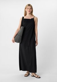 Schwarzes Maxikleid mit Rüschenausschnitt und dünnen Trägern, kombiniert mit einer gestreiften Schwarz-Weiß-Schultertasche und schwarzen flachen Sandalen.