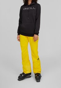 Sudadera negra con capucha y logo blanco, combinada con pantalones de nieve amarillos con bolsillo, y botas de esquí negras con detalles en plata.