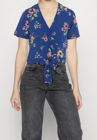 Kvinde iført blå kortærmet blomstret bluse bundet om livet, der er parret med højtaljede blege sorte jeans.