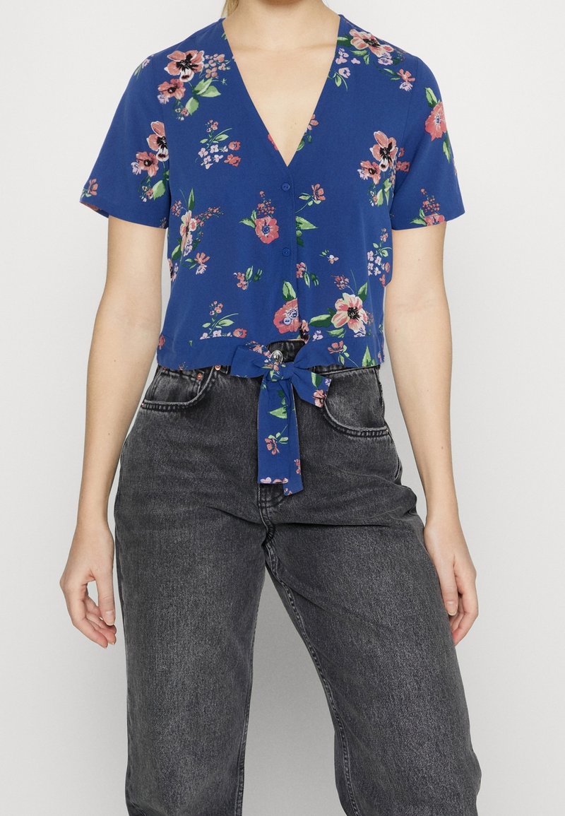 Kvinna i blå blommig blus med kort ärm, knuten i midjan, tillsammans med högmidjade, urblekta svarta jeans.