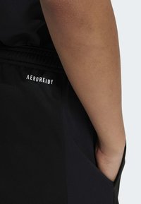 Čierne športové šortky s brandingom AeroReady. Materiál vyzerá hladko s textúrovaným panelom na boku a rukou vo vrecku.
