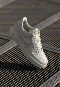 Sapatilhas Nike Air cinza claro com parte superior em couro, caixa de dedo perfurada e atacadores planos. Apresentam sola texturizada e detalhes discretos do logótipo.