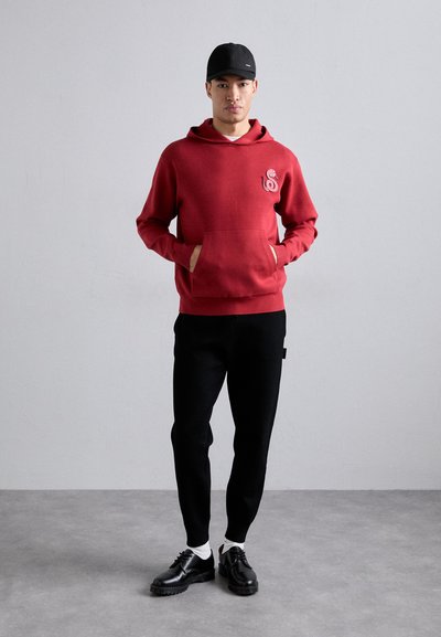Felpa rossa in fleece con tasca frontale e logo, abbinata a pantaloni neri a sigaretta. Scarpe nere con suola spessa e calzini bianchi.