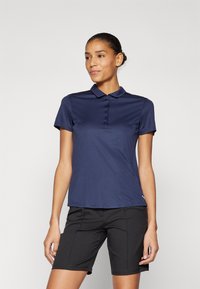 Polo blu navy con maniche corte e colletto, caratterizzato da una chiusura frontale con bottoni e un tessuto liscio e strutturato. Abbinato a pantaloni corti neri.
