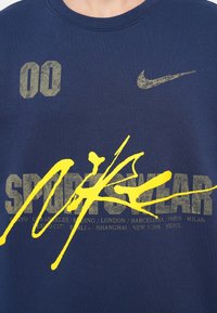 T-maica mornete z modrim odtenkom s velikim rumenim črtnim zamahom in zbledelim sivim besedilom, ki vključuje "00" in "SPORTSWEAR," z logotipom Nike na prsih.