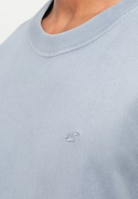 Šviesiai mėlynas medvilninis marškinėliai su apvalia iškirpte, turintys minkštą tekstūrą ir diskretiškai išsiuvinėtą logotipą kairėje krūtinės pusėje.
