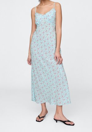 Kobieta w jasnoniebieskiej, kwiatowej sukience na ramiączkach typu spaghetti oraz czarnych sandałach na niskim obcasie, stojąca na tle jednolitego tła.