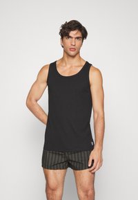 Camiseta sin mangas negra de algodón con cuello redondo, combinada con un bañador a rayas que muestra líneas verticales alternas oscuras y claras.