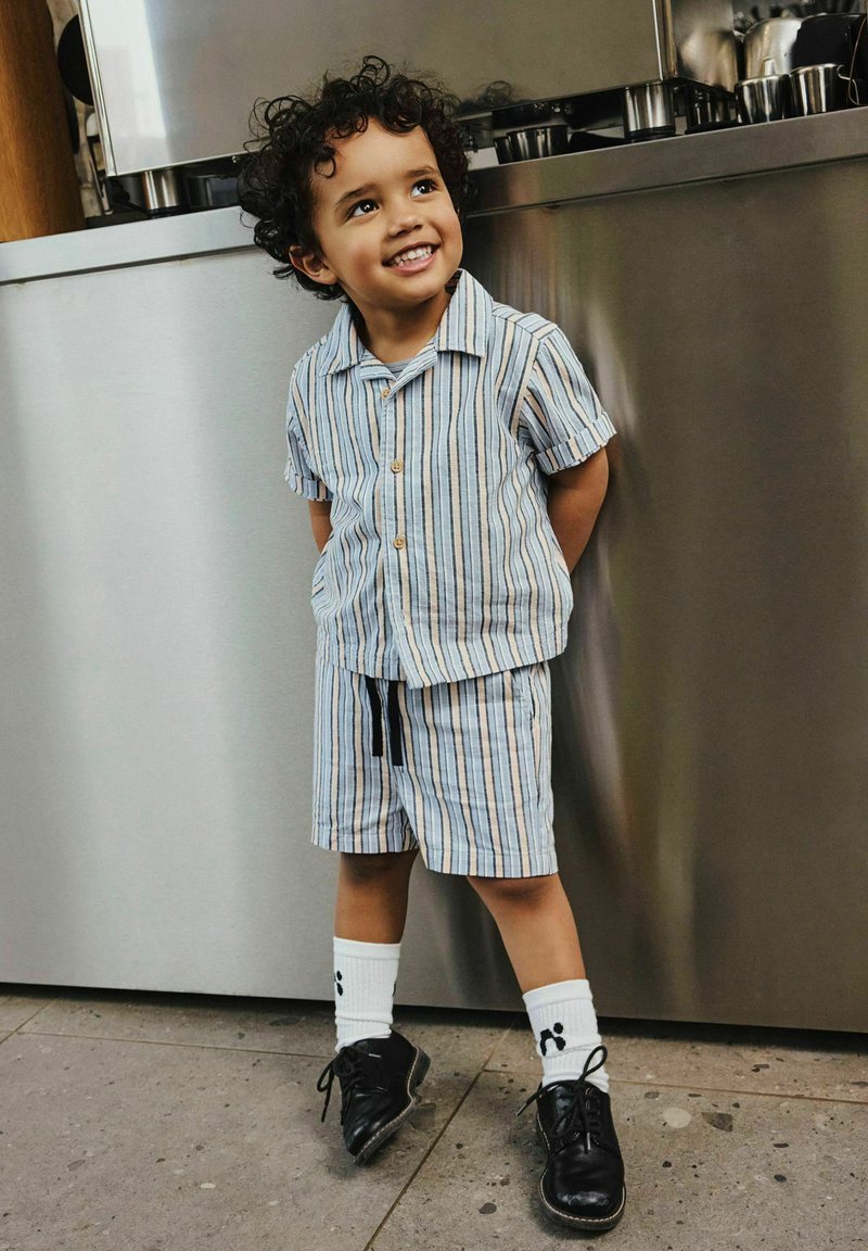 Jeune enfant souriant aux cheveux bouclés, portant une chemise à manches courtes rayée et un short, des chaussettes blanches avec des empreintes de pattes noires, et des chaussures noires à l'intérieur.