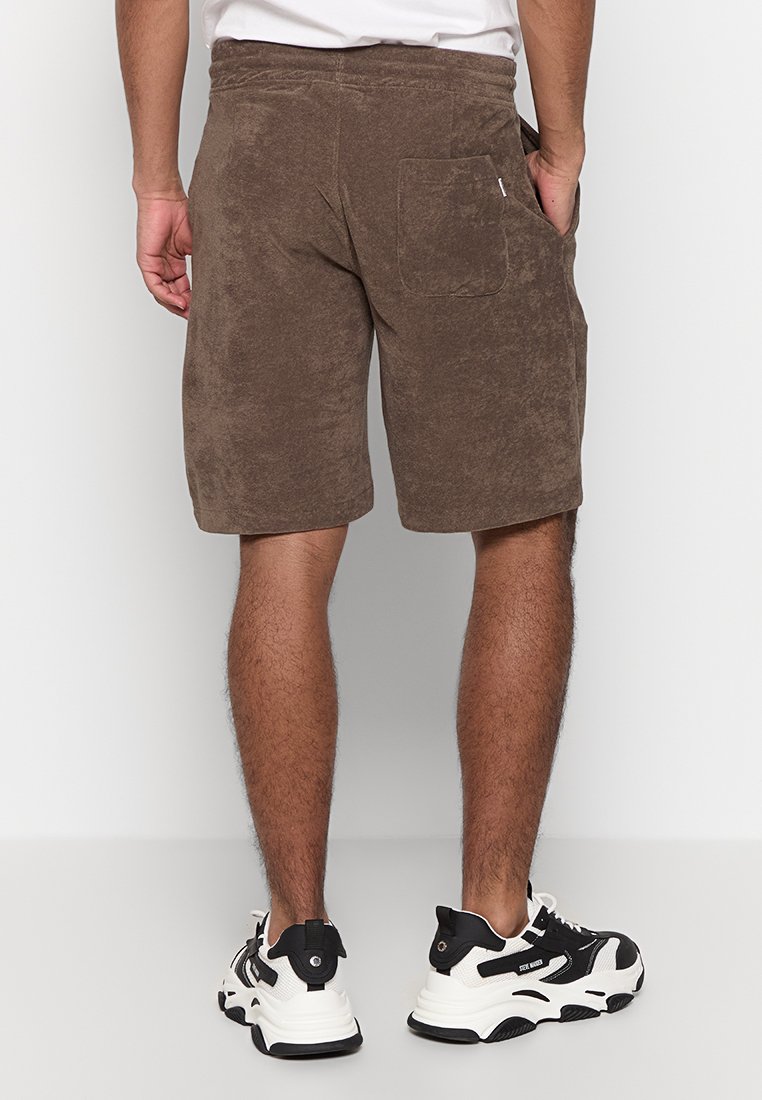 Braune Frottee-Shorts mit einer Gesäßtasche, entspannter Passform und elastischem Bund. Getragen mit schwarz-weißen Sneakers.