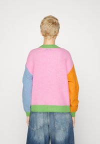 Person mit kurzem blonden Haar, der einen dicken Pullover mit farbigen Blöcken in Pink, Blau, Orange und Grün trägt, kombiniert mit hellblauen Jeans.